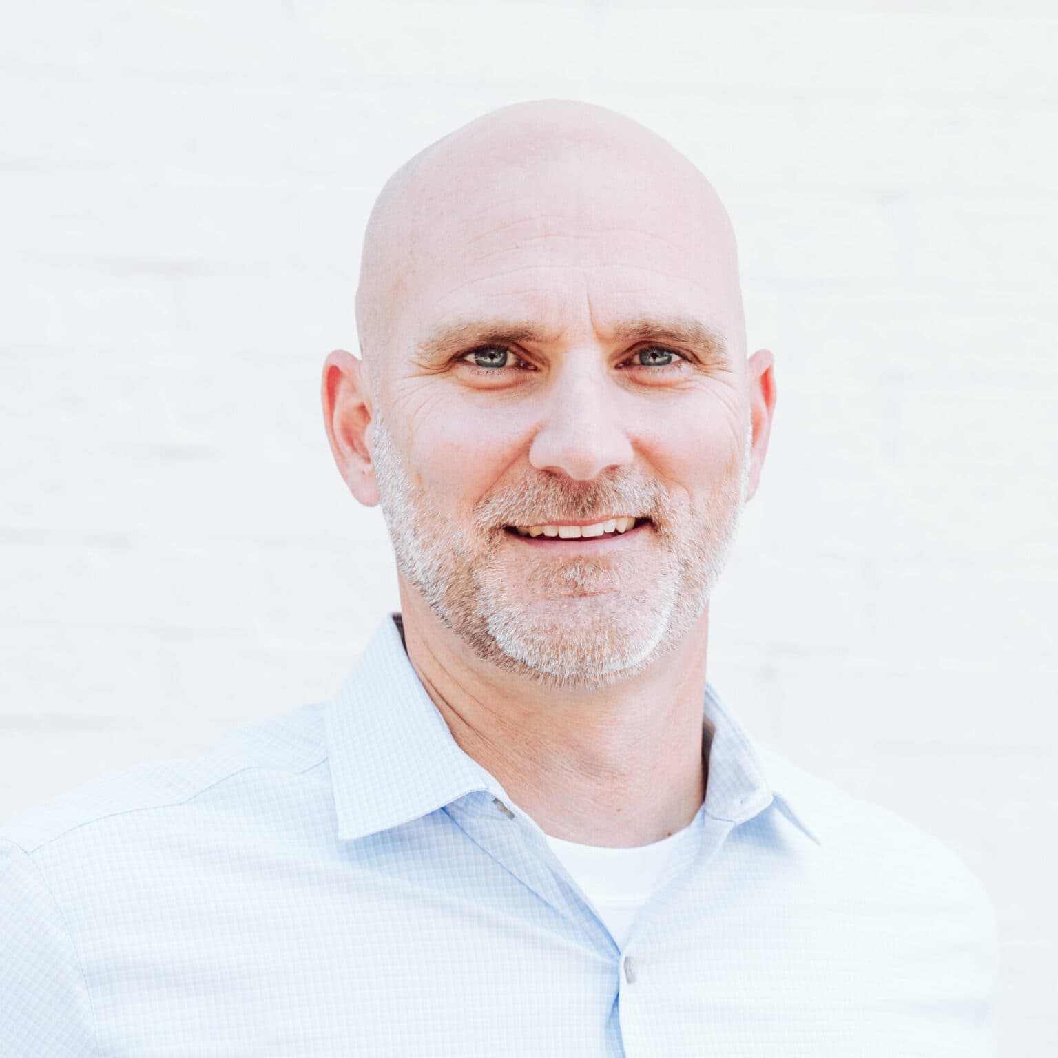 A smiling, middle-aged man with a bald head and trimmed gray beard, wearing a light blue collared shirt, stands in front of a plain white background.